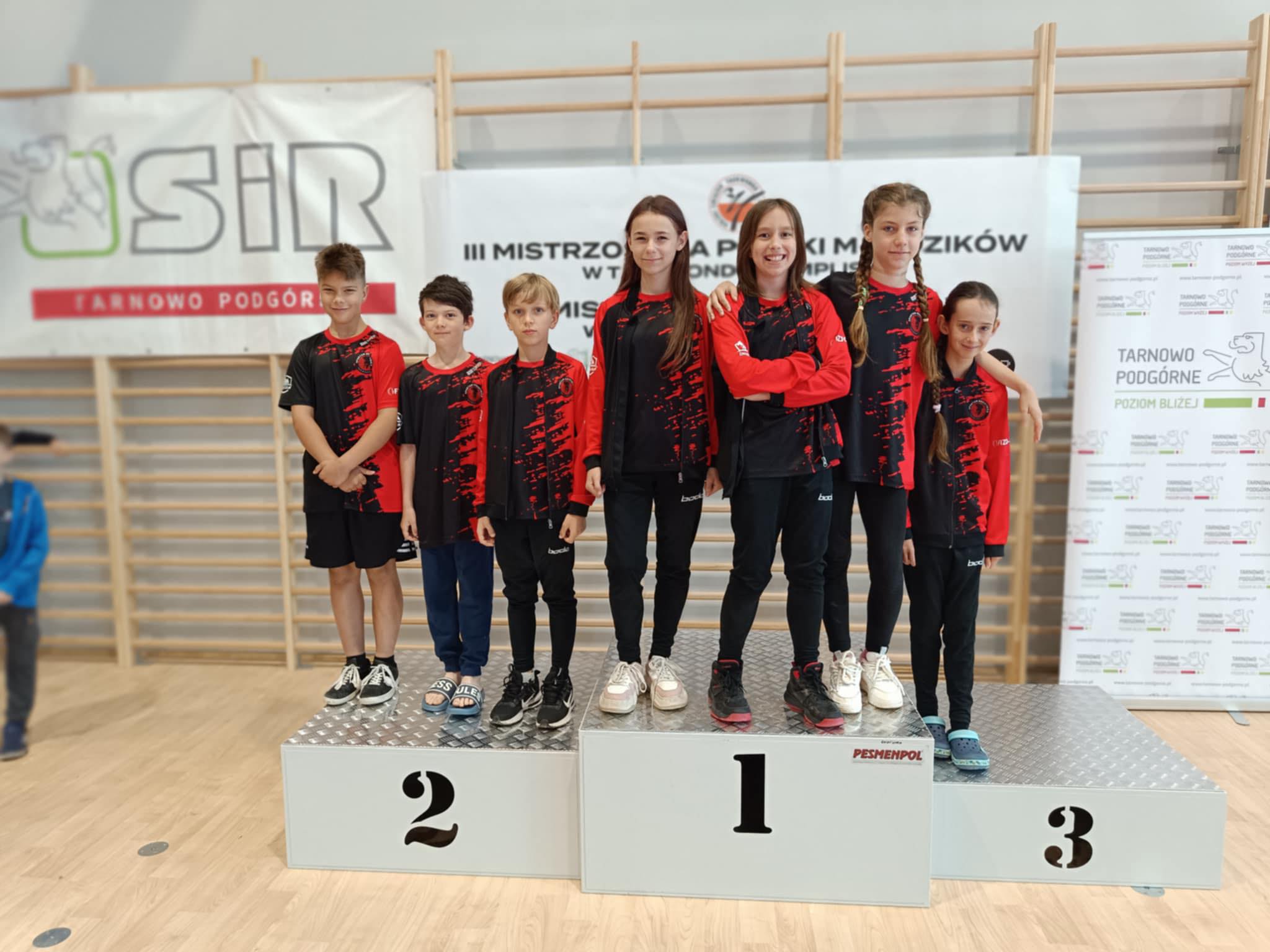 Zawodnicy taekwondo w klubowych strojach stojący na podium.